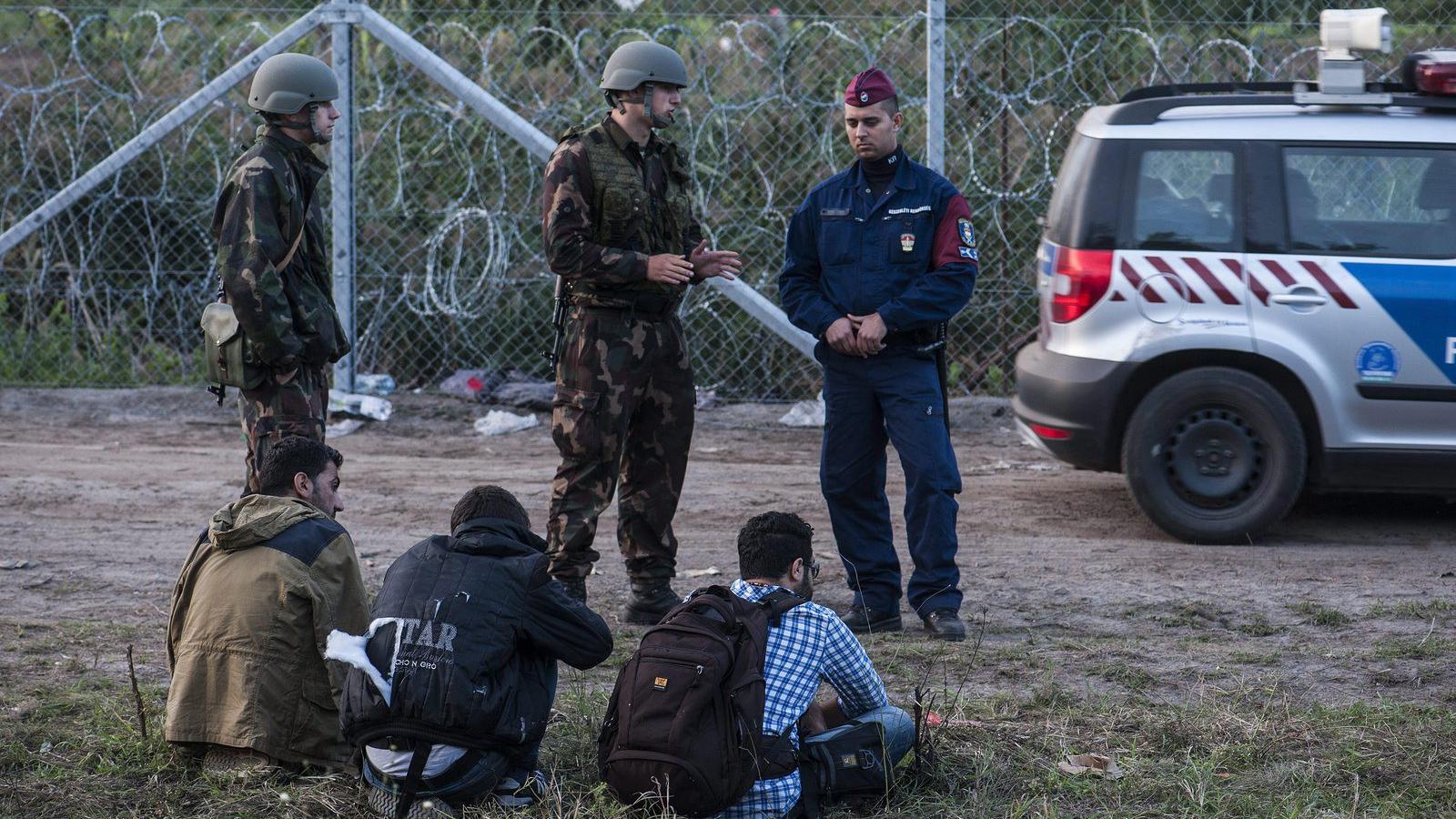 Katonák és rendőrök három elfogott illegális bevándorlót őriznek a biztonsági határzár mellett a szerb-magyar határon, Röszke té